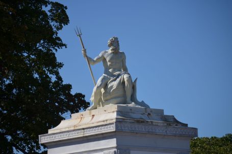 Eine Statue, die eine sitzende Figur mit einem Speer darstellt, vor blauem Himmel.