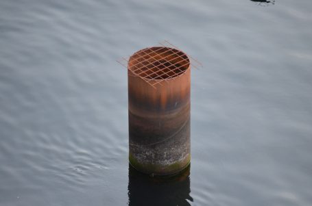 Rostiger, zylindrischer Pfeiler mit Gitterstruktur im Wasser.