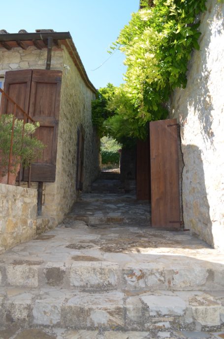 Schmaler gepflasterter Weg zwischen zwei Natursteinmauern mit Holztüren und Pflanzen.
