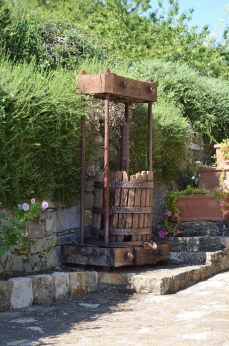Historische Weinpresse aus Holz, umgeben von Grün und Blumen auf einer Steinmauer.