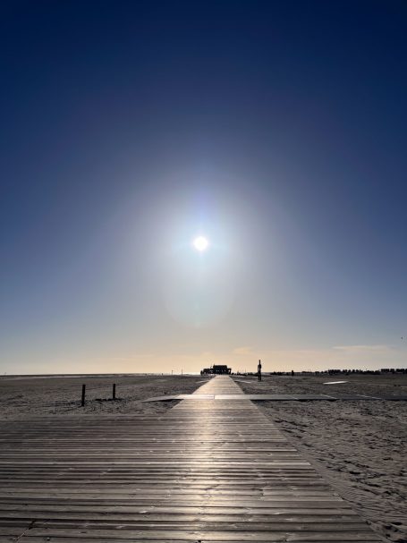 Sonniger Strandweg mit Holzplanken und strahlender Sonne am Horizont.