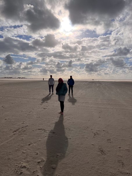 Drei Personen stehen am Strand, umgeben von Wolken und Schatten auf dem Sand.