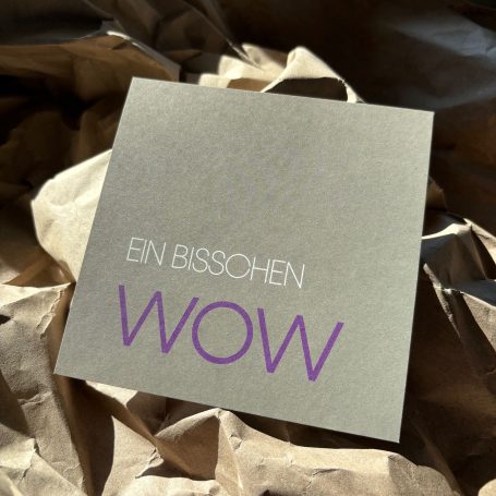 Graue Karte mit dem Text „Ein bisschen WOW“ in lila Schrift.