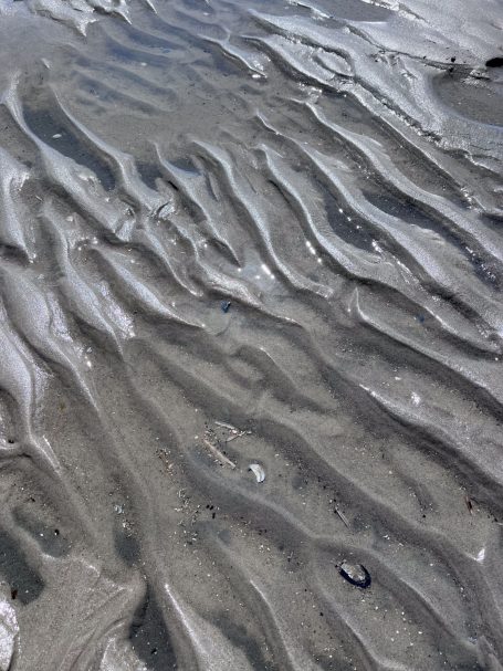 Sandmuster am Strand mit fließenden Wellen und kleinen Muscheln.