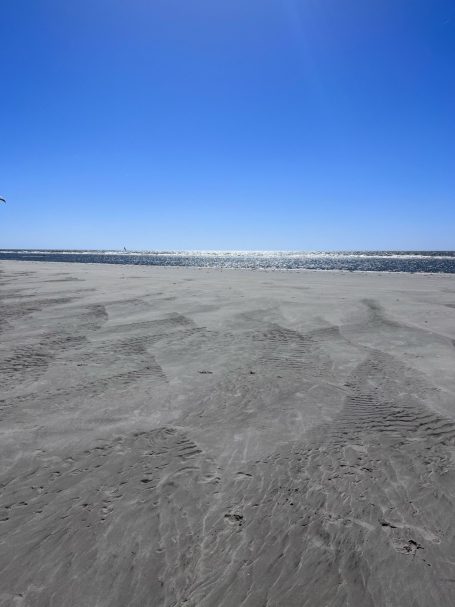 Weitläufiger Sandstrand unter klarem blauen Himmel mit sanften Wellen im Hintergrund.