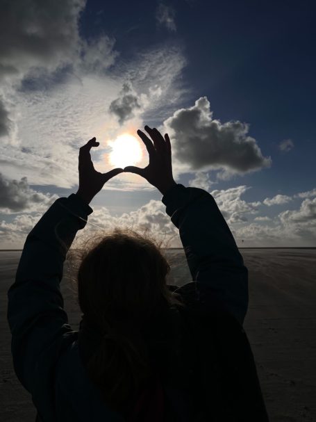 Eine Person hält die Hände in Form eines Rahmens vor die Sonne mit Wolken im Hintergrund.